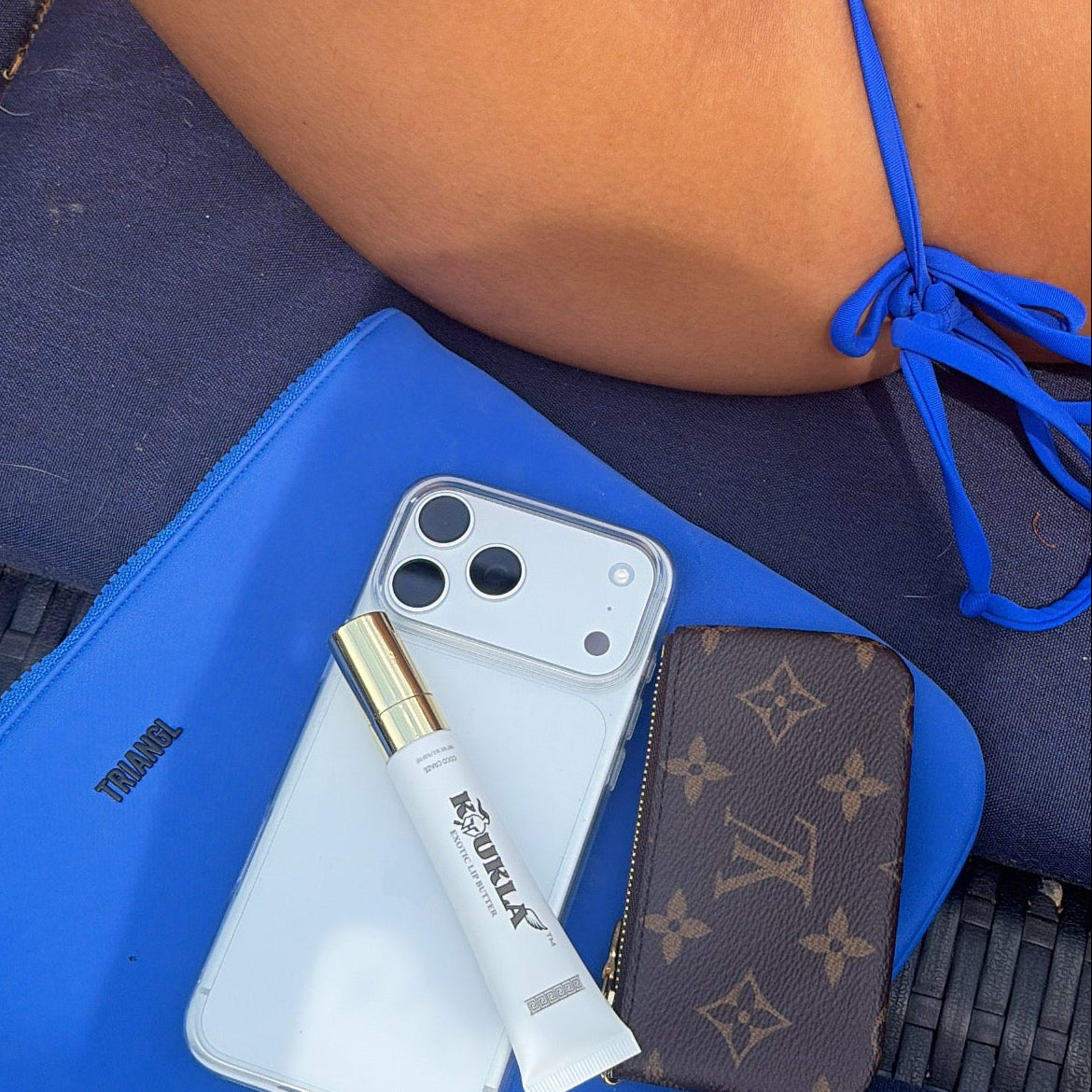 Phone, lip butter, and wallet on a blue surface with a person wearing a blue bikini in the background.