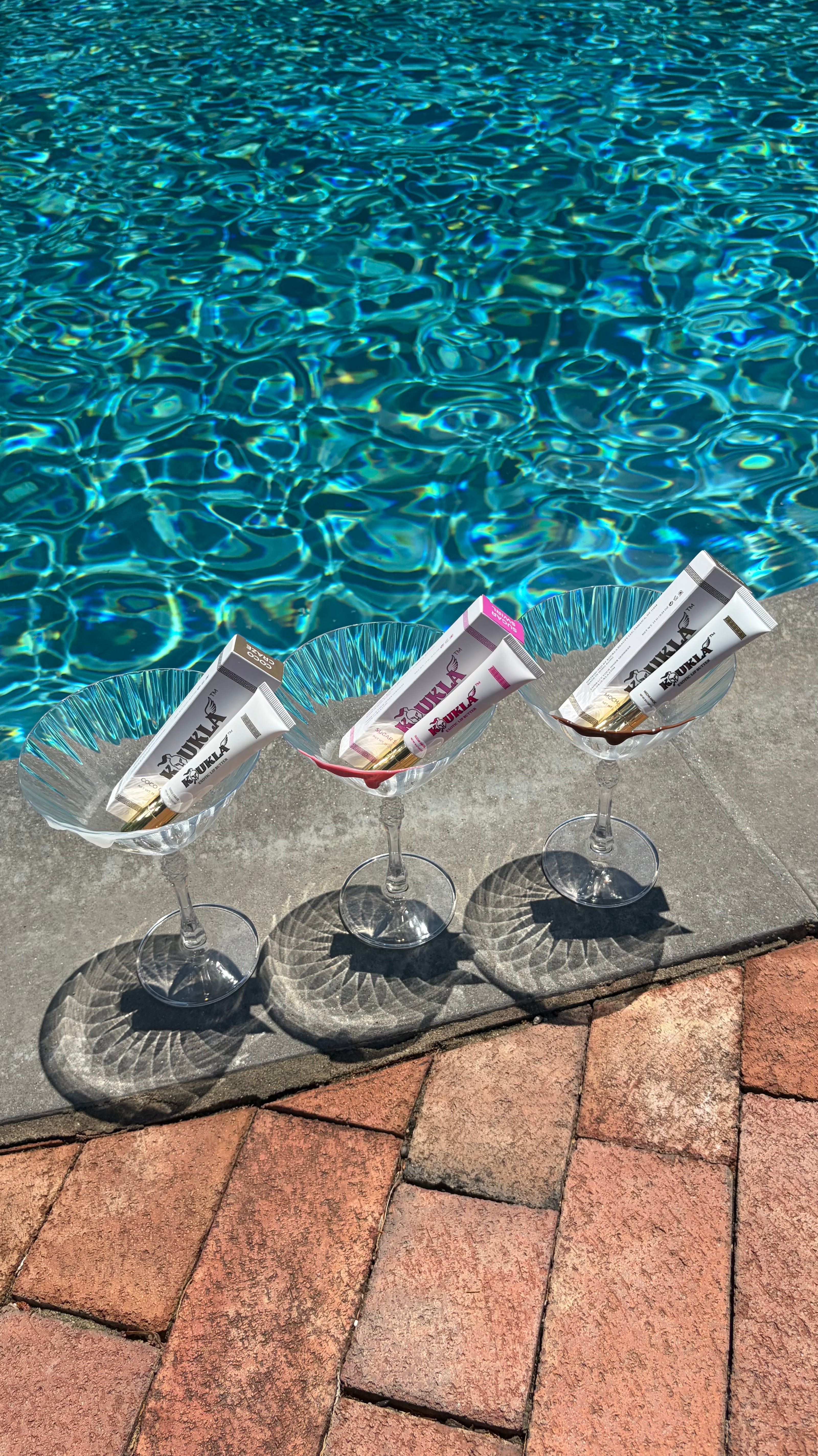 Martinis with lip butter tubes labeled "Koukla"resting in clear stemmed cocktail glasses on a concrete poolside ledge, with bright blue swimming pool water in the background and red brick pavers in the foreground.