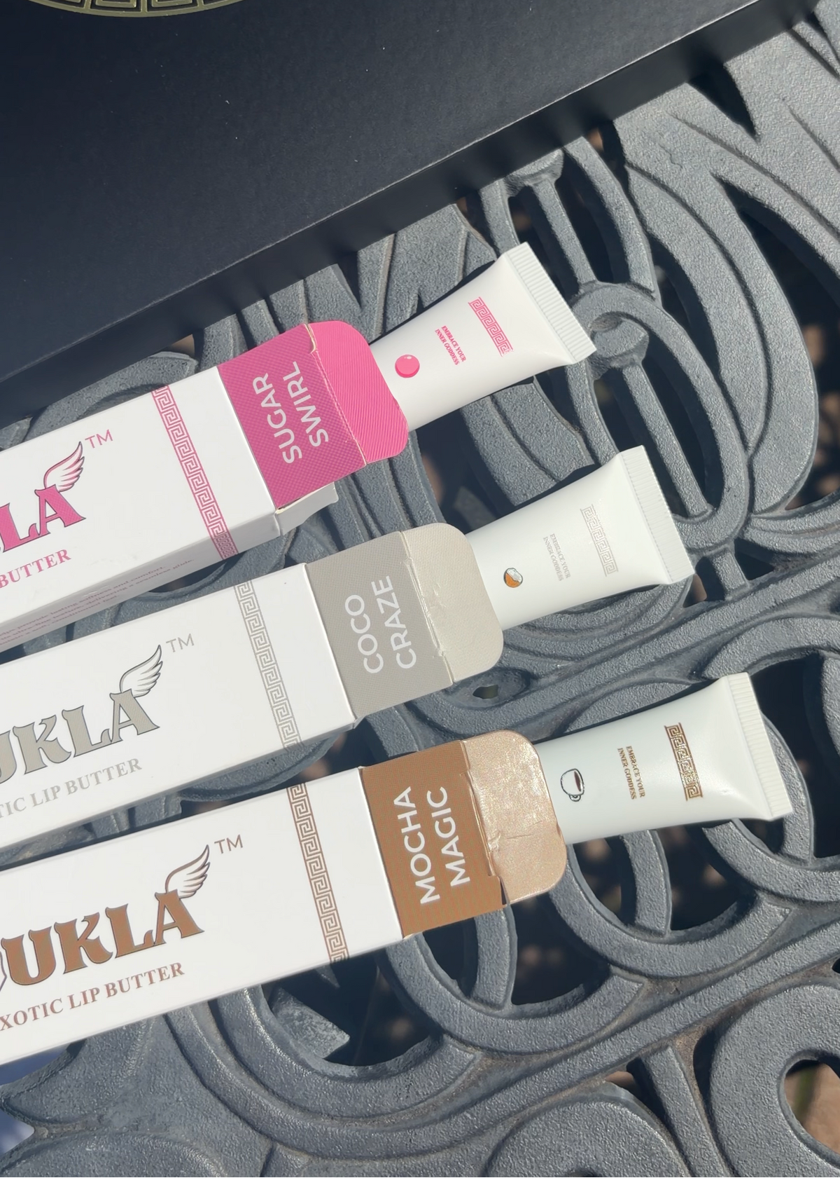 Three tubes of Koukla lip butter with their matching boxes arranged diagonally on a dark metal patio chair. The lip butters are in Sugar Swirl, Coco Craze, and Mocha Magic flavors.