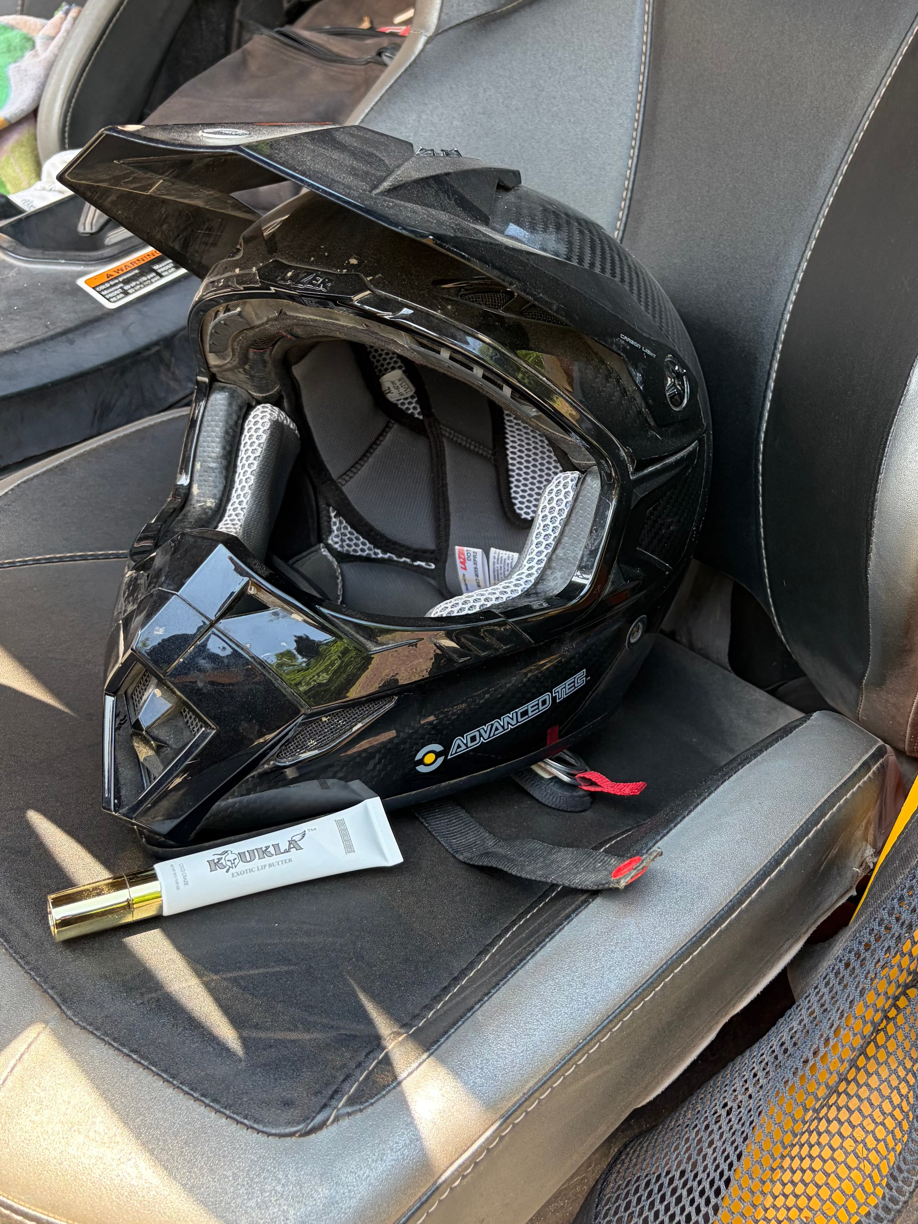 A black full-face motorcycle helmet with its visor open, resting on a gray seat inside a vehicle. A tube of Koukla Lip Butter is placed on the seat next to the helmet.